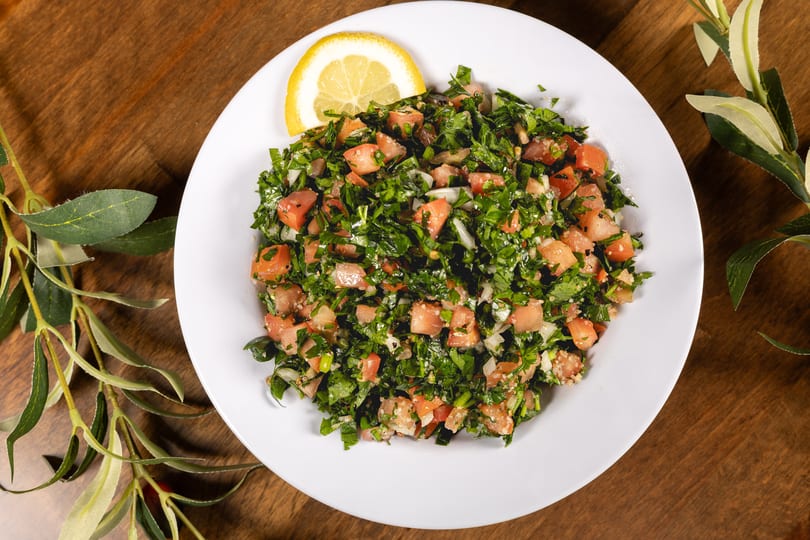 A vibrant tabbouleh salad with chopped parsley, tomatoes, and a lemon slice on a white plate.