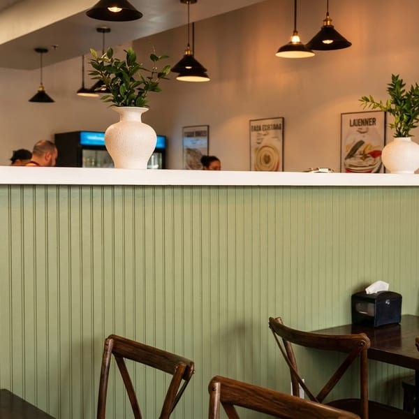 Restaurant interior with a green paneled counter, dark wooden tables, chairs, and pendant lights.