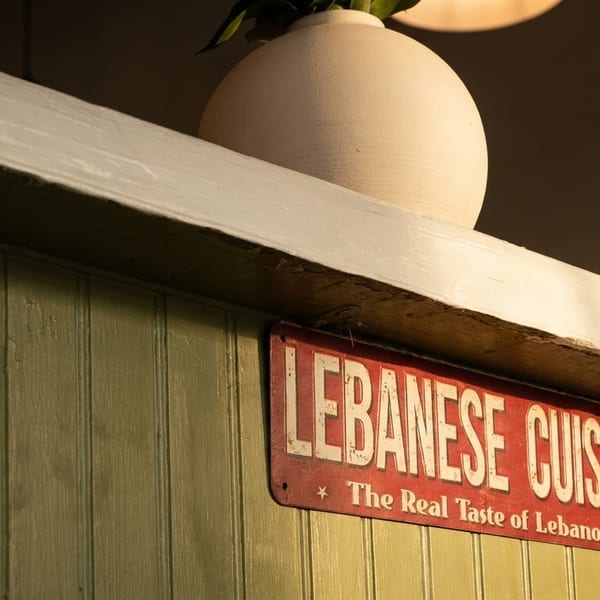 A red sign with "LEBANESE CUISINE" in white hangs on a green wooden wall, with a white vase above.